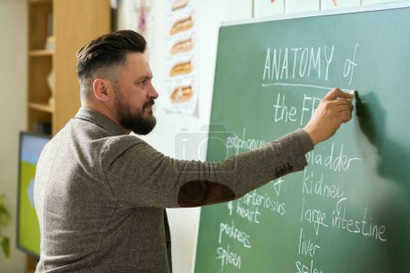 Photo for Mature teacher writing down names of frog internal organs on blackboard while explaining students of high school subject of lesson - Royalty Free Image