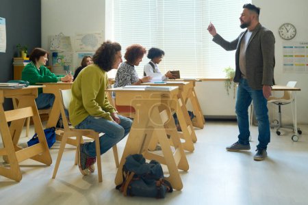 Photo for Group of students sitting by desks in classroom and listening to explanation of teacher standing in front of them at seminar - Royalty Free Image