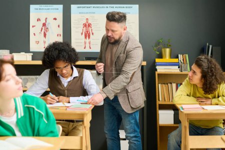 Photo for Mature male teacher of anatomy checking notes of highschool student and consulting him among classmates at lesson in classroom - Royalty Free Image