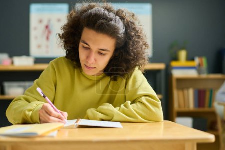 Téléchargez les photos : Adolescent mâle en pull jaune prendre des notes dans son copybook tout en étant assis par bureau à la leçon et l'exécution d'une mission individuelle - en image libre de droit