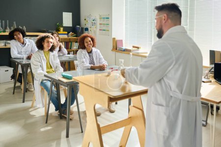 Photo for Teacher of chemistry showing experiment to group of students at lesson while standing by desk and mixing chemical substances - Royalty Free Image