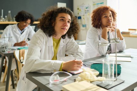 Photo for Group of clever intercultural students of highschool in labcoats listening to explanation of their teacher while sitting by desk at chemistry - Royalty Free Image