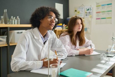 Téléchargez les photos : Deux adolescents intelligents en blouse de laboratoire regardant le professeur hteir assis près des bureaux à la leçon de chimie et l'écoutant - en image libre de droit