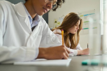 Photo for Youthful girl in labcoat making notes in copybook during laboratory work at lesson of chemistry while sitting next to her classmate - Royalty Free Image