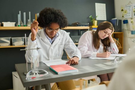 Photo for Two clever intercultural teenage students in labcoats sitting by desk and carrying out individual assignment at lesson of chemistry - Royalty Free Image