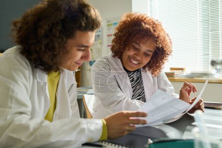 Photo for Teenage guy in labcoat showing paper with test tasks to African American female classmate sitting next to him at lesson of chemistry - Royalty Free Image
