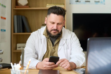 Photo for Serious mature teacher in labcoat scrolling in smartphone or texting while sitting by workplace against wooden shelves with books - Royalty Free Image