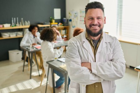 Photo for Happy bearded mature teacher of chemistry in labcoat crossing his arms on chest and looking at camera against group of teenage students - Royalty Free Image