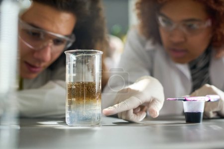 Photo for Teenage student in protective gloves pointing at glass containing chemical dissolver during experiment at lesson of chemistry - Royalty Free Image