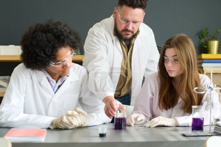 Foto de Profesora segura de química tomando vidrio con mezcla púrpura de sustancias químicas mientras ayuda a los estudiantes con el experimento - Imagen libre de derechos