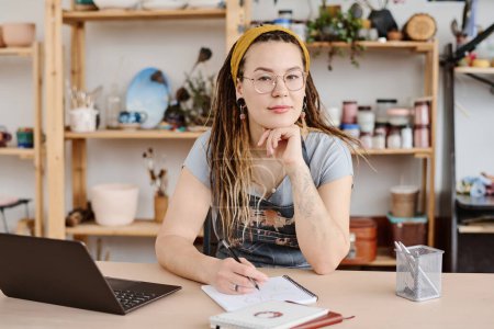 Téléchargez les photos : Jeune femme moderne en tenue décontractée et lunettes assis par le lieu de travail en studio et regardant la caméra tout en faisant un croquis dans le bloc-notes - en image libre de droit