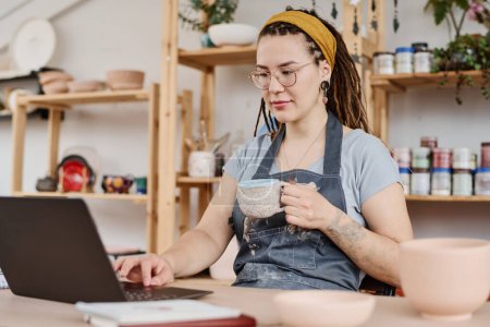 Téléchargez les photos : Jeune femme pigiste ou propriétaire d'une petite entreprise assise devant un ordinateur portable par lieu de travail, prenant le thé et à la recherche de nouvelles idées dans le net - en image libre de droit