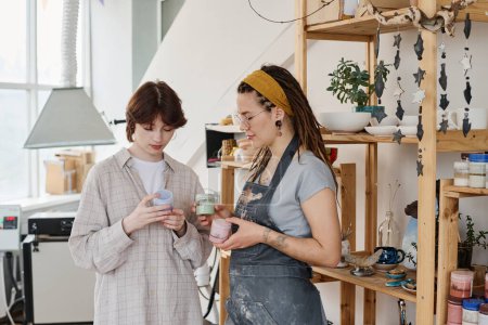 Foto de Joven cliente femenino eligiendo gouache para pintar artículos de barro hechos a mano y consultando con el empleado que trabaja en la tienda - Imagen libre de derechos