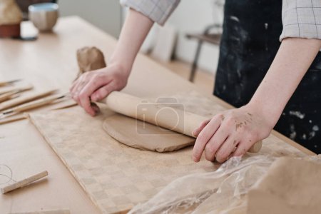 Téléchargez les photos : Mains de jeune artisan avec rouleau en bois aplatissant l'argile à bord tout en travaillant sur de nouveaux produits en argile à vendre en atelier - en image libre de droit