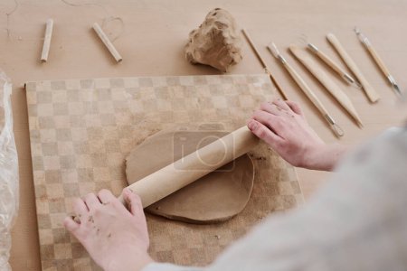 Téléchargez les photos : Gros plan des mains de jeunes femmes créatives aplatissant l'argile à bord avec rouleau à pâtisserie tout en créant un nouvel article en faïence dans l'atelier - en image libre de droit