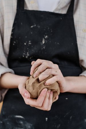 Téléchargez les photos : Mains de jeune artisan femelle en tablier noir pétrissant un morceau d'argile pour la fabrication de faïence ou d'autres objets créatifs en argile à vendre - en image libre de droit