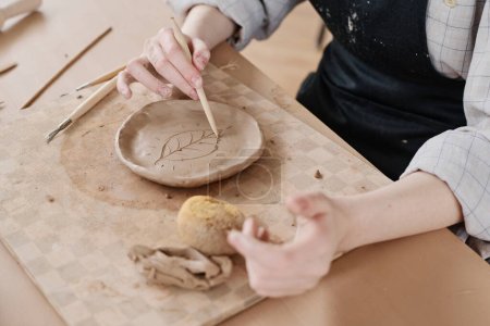 Téléchargez les photos : Mains de la jeune femme maître poterie feuille de coupe sur plaque d'argile pièce à travailler à bord tout en étant assis à table dans l'atelier - en image libre de droit