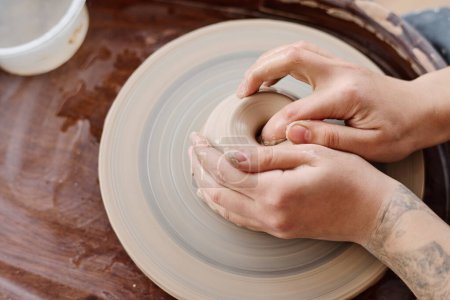 Téléchargez les photos : Plan ci-dessus des mains d'une jeune potière créative ou propriétaire d'un atelier assis par une roue de poterie et fabriquant un produit en argile - en image libre de droit