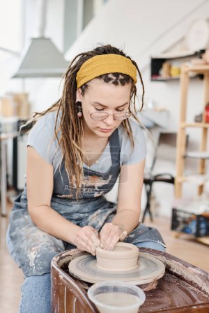 Téléchargez les photos : Femme créative en tenue décontractée assise en tournant la roue de poterie en atelier et en créant de nouveaux articles en argile pour la vente en faïence - en image libre de droit