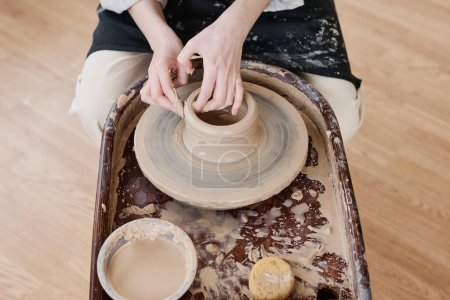 Téléchargez les photos : Mains de jeune artisan assis en tournant la roue de poterie dans l'atelier et en créant un pot ou une cruche en argile à vendre dans un magasin de faïence - en image libre de droit
