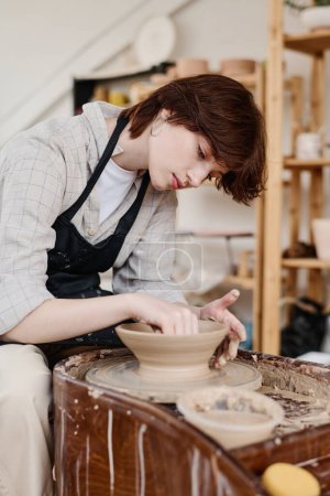 Téléchargez les photos : Jeune lanceuse se penchant sur la roue de poterie rotative pendant le travail sur un nouvel objet en argile alors qu'elle était assise dans son atelier ou son studio - en image libre de droit