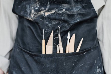 Foto de Parte de artesano en delantal negro con herramientas necesarias para tallar adornos en artículos de arcilla hechos a mano en el bolsillo delantero - Imagen libre de derechos