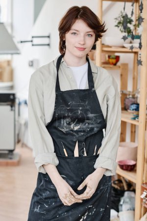 Photo for Young creative female pitcher in workwear standing in front of camera against interior of workshop or studio and wooden shelves - Royalty Free Image