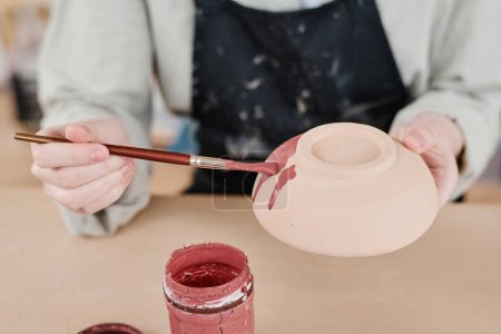 Téléchargez les photos : Jeune artisan créative féminine avec pinceau colorant bol en terre cuite à la main en marsala tout en travaillant par table en atelier - en image libre de droit