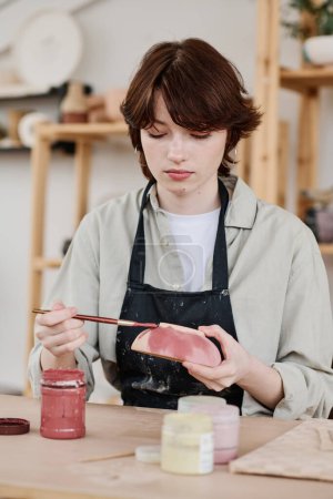 Téléchargez les photos : Jeune artisan féminin qualifié sérieux avec bol en argile et peinture au pinceau faïence faite à la main tout en étant assis près du lieu de travail - en image libre de droit