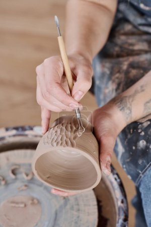 Téléchargez les photos : Mains de jeunes céramistes féminines créatives décorant à la main tasse en argile avec des motifs sculptés en atelier tout en utilisant un outil à main pointu - en image libre de droit