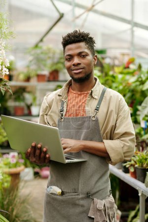 Téléchargez les photos : Portrait de jeune homme africain en uniforme tenant un ordinateur portable dans les mains et regardant la caméra debout dans la serre - en image libre de droit