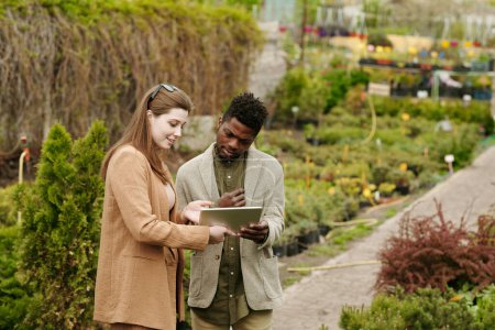 Téléchargez les photos : Jeune femme montrant son projet de maison sur tablette numérique au paysagiste pendant qu'ils achètent des plantes dans le jardin - en image libre de droit