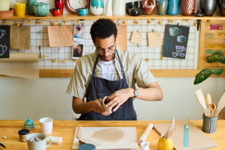 Téléchargez les photos : Jeune homme noir créatif en vêtements de travail créant un nouvel article en argile à vendre assis à table contre une étagère avec de la faïence en atelier - en image libre de droit