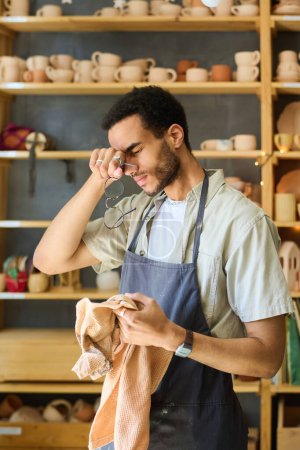 Photo for Young tired black man with towel touching his head after hard work over creating new handmade clay items for sale in workshop - Royalty Free Image