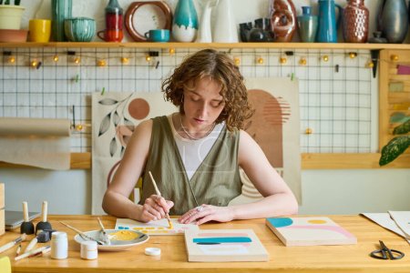 Téléchargez les photos : Jeune femme créative en tenue décontractée avec pinceau et gouache créant une nouvelle peinture tout en étant assise sur le lieu de travail en studio - en image libre de droit
