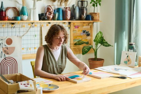 Téléchargez les photos : Jeune femme créative avec pochoir en papier créant de la peinture abstraite tout en étant assis à table en studio avec diverses fournitures pour les œuvres d'art - en image libre de droit