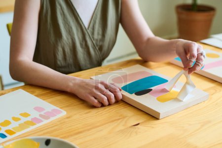 Téléchargez les photos : Mains de jeune femme créative assise à côté d'une table en bois dans un atelier ou un studio et utilisant un pochoir tout en créant une nouvelle peinture abstraite - en image libre de droit