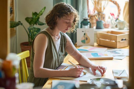 Téléchargez les photos : Jeune femme sérieuse avec pinceau assis sur le lieu de travail en atelier et peignant des choses abstraites avec des aquarelles ou de la gouache sur papier - en image libre de droit