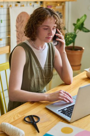Photo for Young serious woman with smartphone by ear talking to client while sitting by table in front of laptop and looking through online orders - Royalty Free Image