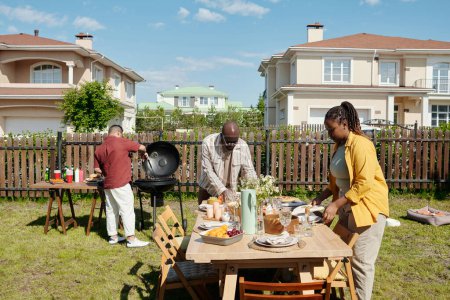 Photo for Young black man and woman serving table with homemade food, drinks, wildflowers and other stuff for outdoor party on summer day - Royalty Free Image