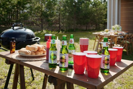 Photo for Wooden table with homemade food, bottles of beer, plastic cups, ketchup prepared for barbeque party standing in front of country house - Royalty Free Image