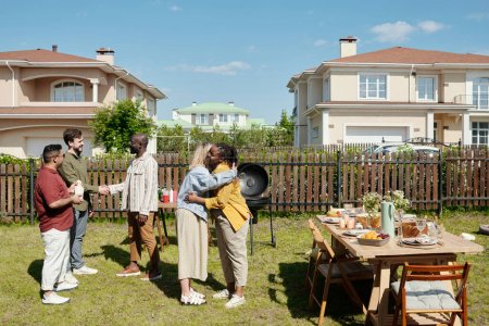 Téléchargez les photos : Heureux jeune homme et femme noirs rencontrer des invités par table servie préparé pour la fête en plein air par leur maison de campagne le week-end d'été - en image libre de droit