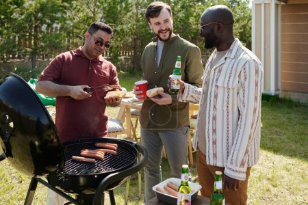 Foto de Tres jóvenes y felices hombres interculturales tomando cerveza y charlando durante la fiesta al aire libre el fin de semana mientras uno de ellos hace una barbacoa - Imagen libre de derechos