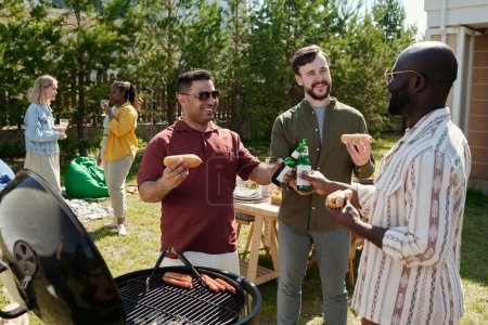Photo for Group of young cheerful intercultural men clinking with bottles of beer during outdoor party against served table and two girls - Royalty Free Image