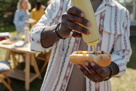 Foto de Manos de joven negro poniendo mostaza en la parte superior de hot dog con salchicha a la parrilla mientras se prepara bocadillo durante la fiesta al aire libre - Imagen libre de derechos