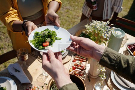 Foto de Manos de hombre joven tomando plato con ensalada de verduras que pasa por la mujer negra sobre la mesa servida con comida y bebidas caseras - Imagen libre de derechos