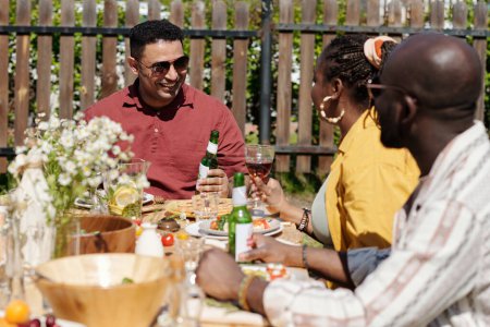 Foto de Joven pareja negra y hombre hispano con copas de vino y botellas de cerveza charlando durante el picnic o fiesta al aire libre por mesa servida - Imagen libre de derechos