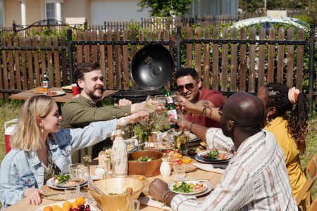 Foto de Grupo de jóvenes amigos interracial con bebidas tostadas durante la cena al aire libre o fiesta sobre la mesa servida por comida y bebidas caseras - Imagen libre de derechos