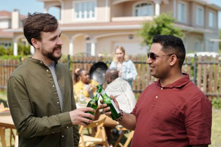 Foto de Feliz joven interracial amigos tintineo con botellas de cerveza y mirando el uno al otro mientras disfruta de la reunión al aire libre - Imagen libre de derechos