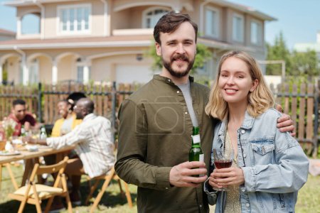 Photo for Happy young couple with alcoholic drinks looking at camera while enjoying outdoor gathering against group of intercultural friends - Royalty Free Image